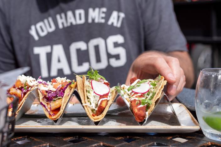Four tacos resting in a holder with a person behind wearing a shirt that says 'you had me at tacos'